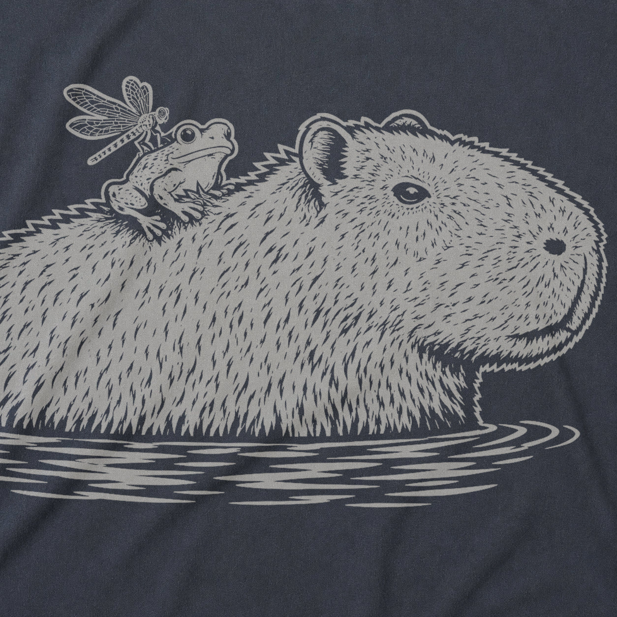 A close-up of a dark navy blue t-shirt featuring a detailed graphic print of a capybara in water with a frog sitting on its back and a dragonfly above it, all in a light gray sketch style.